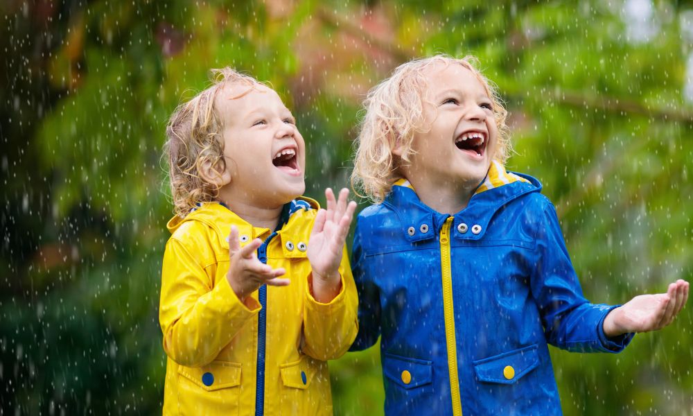 two kids in raincoats look up at the rain in the sky and laugh
